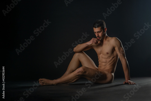 Fashion nude photo of a male model with seductive figure sitting isolated on the floor in a studio