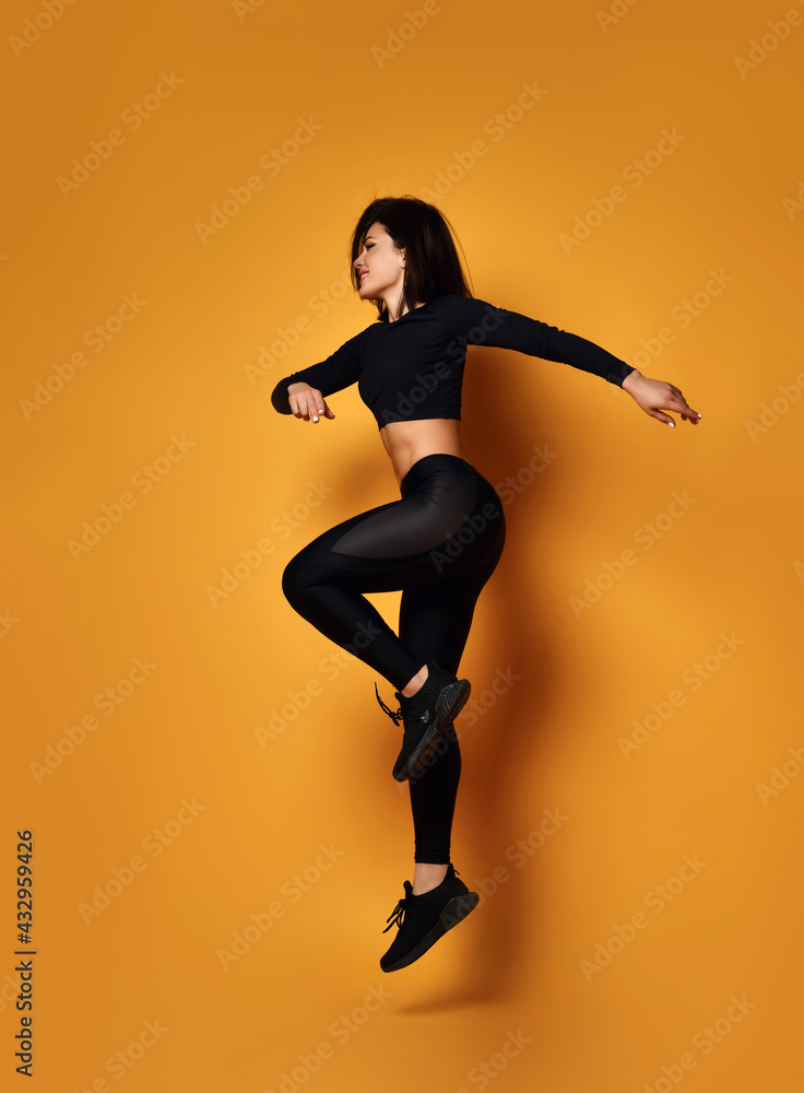 Young smiling sexy pretty lady in black sportswear jumping during workout over yellow background, side view. Beautiful women and active sport lifestyle concept Young smiling sexy pretty lady in black sportswear jumping during workout over yellow background, side view. Beautiful women and active sport lifestyle concept