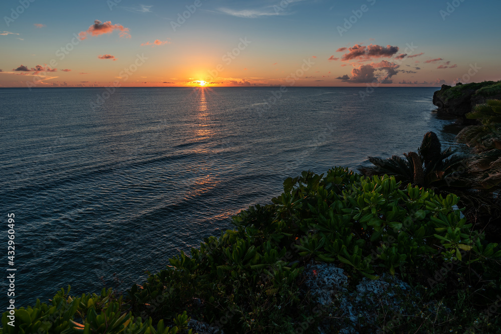 Amazing sunset with its orange rays descending on the horizon reflecting in the sea. Blue sky, pink clouds. Sea with a crisp surface due to the gentle coastal wind, seaside plant composing the scene.
