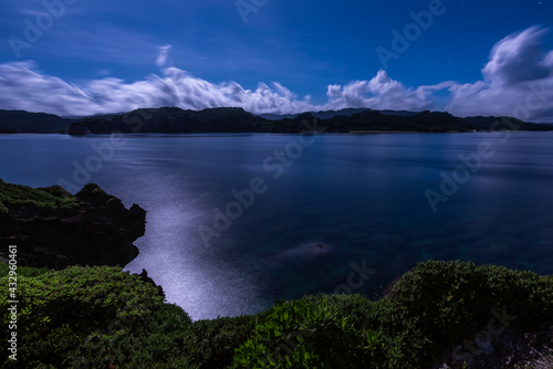 Contemplative scene of a long exposure under the moonlight. Flat sea with reflection of moon light, mountains, clouds in blurred motion, trail of stars. Vegetation in foreground.