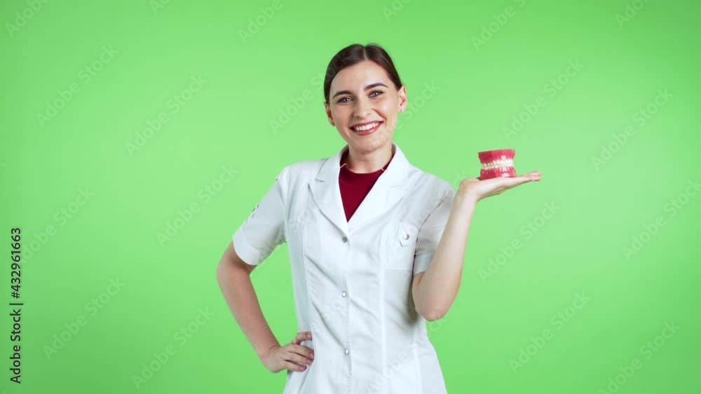 Cheerful attractive female dentist is holding jaws on her hand, looking into the camera and smiling. Green screen is on the background Cheerful attractive female dentist is holding jaws on her hand, looking into the camera and smiling. Green screen is on the background