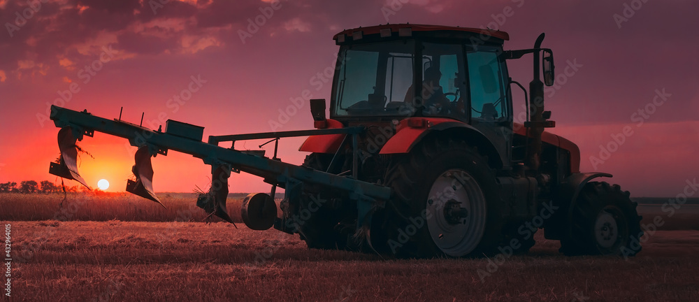 Fototapeta premium Tractor with a raised plow in an agricultural field against the backdrop of a sunset. Plowing after harvest. Concept, agriculture, food industry. Selective focus, toned photograph.