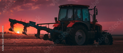 Tractor with a raised plow in an agricultural field against the backdrop of a sunset. Plowing after harvest. Concept, agriculture, food industry. Selective focus, toned photograph.