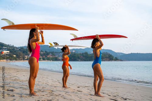 Portrait group of Asian woman girl friends in swimwear holding surfboard standing on the beach together at summer sunset. Female friendship enjoy outdoor activity lifestyle play extreme sports surfing