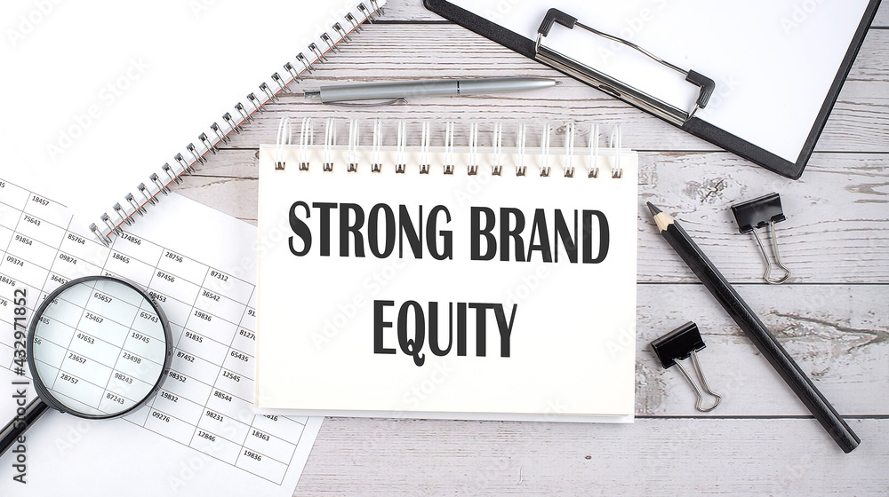 Notepad with text STRONG BRAND EQUITY with a pen,office tools and graphs on the desktop. Notepad with text STRONG BRAND EQUITY with a pen,office tools and graphs on the desktop.