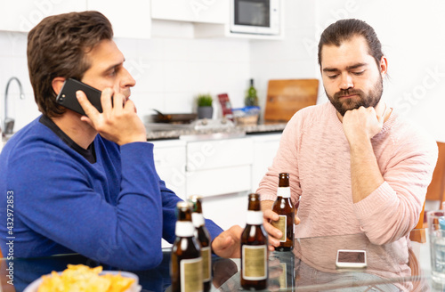 Two concerned men talking and laughing while enjoying beer at home, using phone. High quality photo