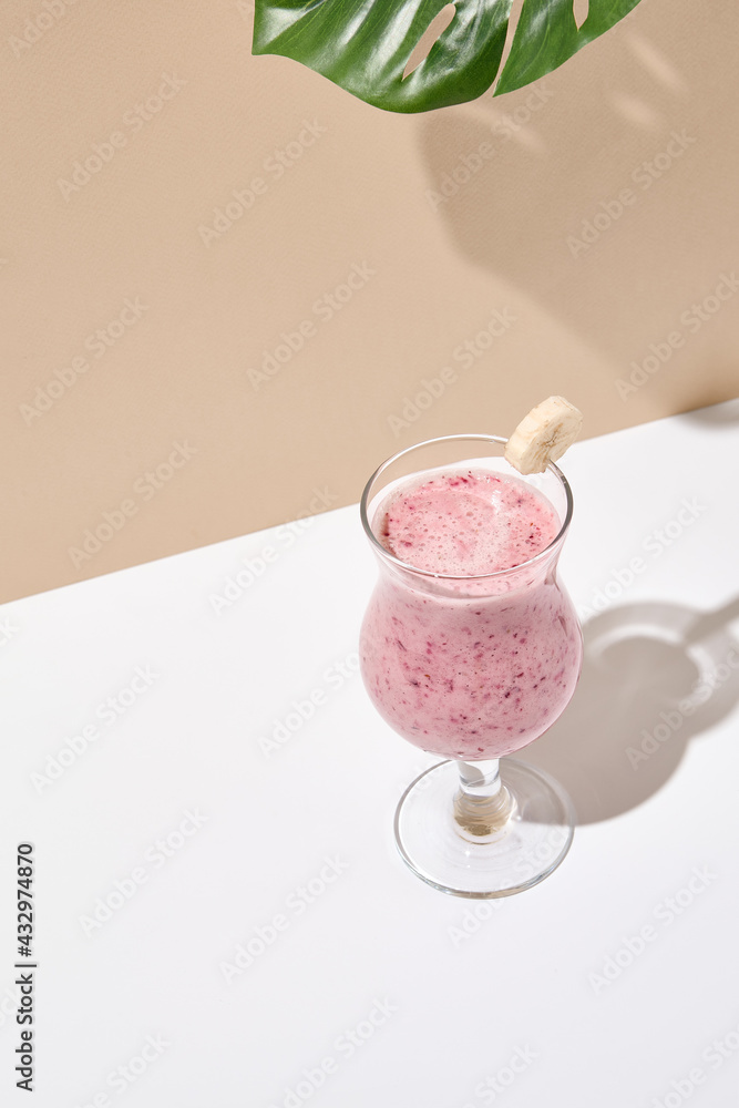 Berry smoothie with banana and milk on white table. Summer healthy drink concept. Beige wall and white table with sunshine and palm leaf shadow. - obrazy, fototapety, plakaty 