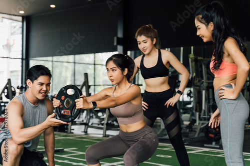 Group of people cheering on their Asian female friend doing squats with a weight plate in fitness gym. Working out together as a teamwork. Encouragement and togetherness concept