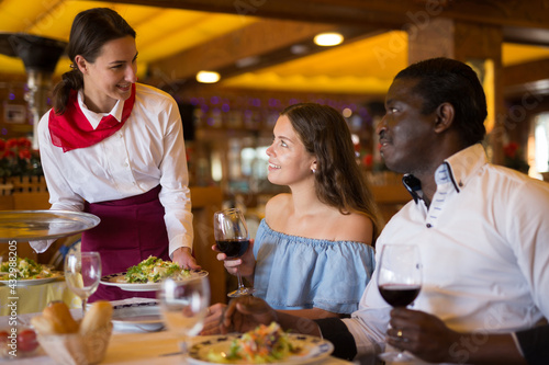Hospitable young beautiful waitress  bringing dishes to couple of guests at restaurant