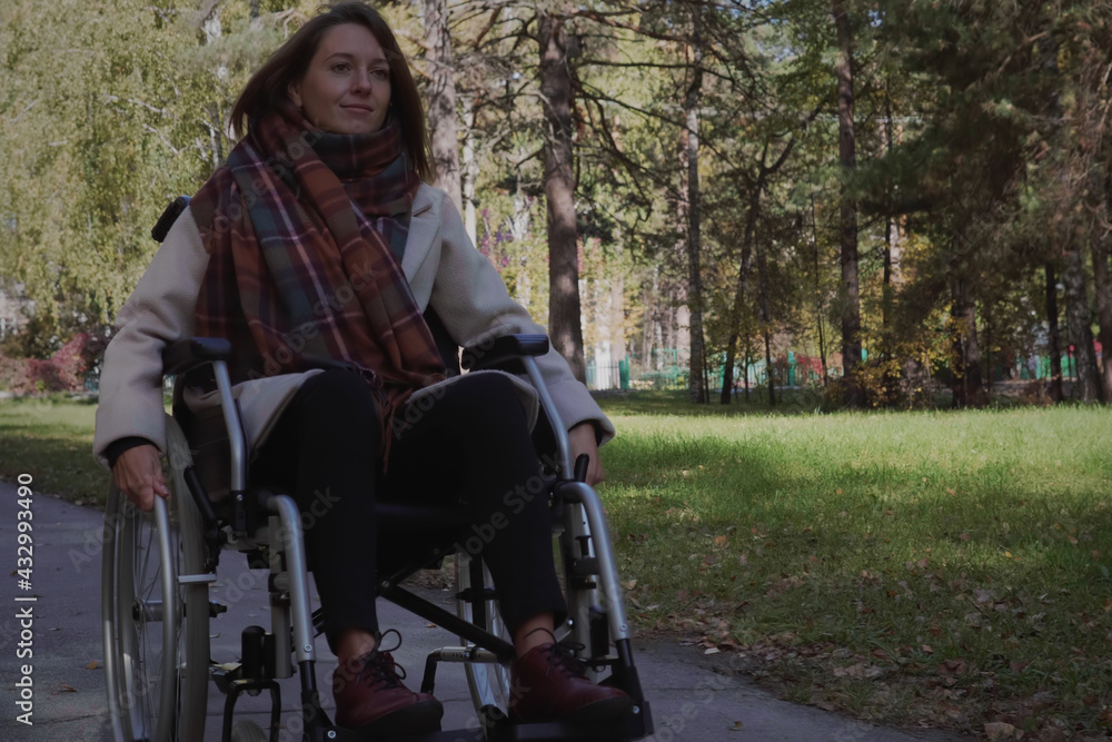 Beautiful brunette woman wearing warm scarf walking on a wheelchair in the autumn city park.