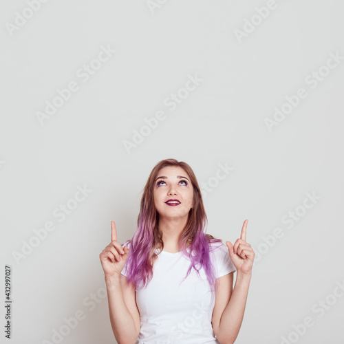 Young attractive woman in white casual t shirt looking and pointing up with both index fingers, copy space for advertisement or promotional text, posing isolated over gray background.