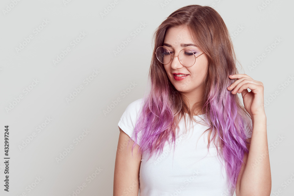 Dreamy attractive woman with lilac hair looking away and touching her hair, wearing white t shirt and glasses, copy space for advertisement, posing isolated over gray background. Dreamy attractive woman with lilac hair looking away and touching her hair, wearing white t shirt and glasses, copy space for advertisement, posing isolated over gray background.