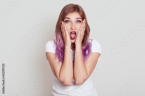 Winsome lady with lilac hair expressing astonishment, sees something surprising, hears shocking news, keeps mouth opened, holds palms on cheeks, posing isolated over gray background.