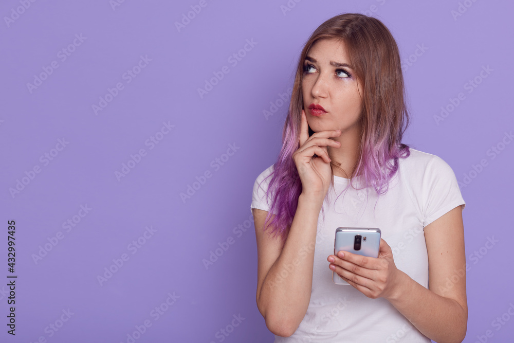 Young adorable thoughtful woman holding smart phone in hands, looking away with pensive expression, keeping finger on cheek, thinks, copy space, posing isolated over purple background. Young adorable thoughtful woman holding smart phone in hands, looking away with pensive expression, keeping finger on cheek, thinks, copy space, posing isolated over purple background.
