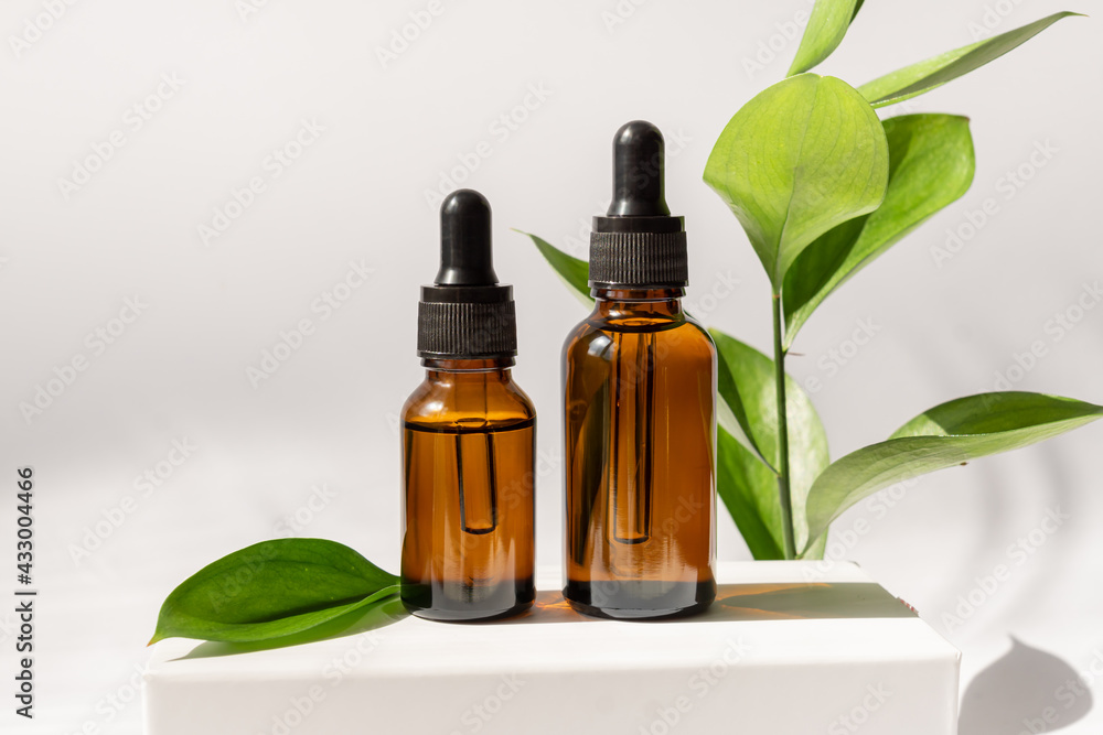 Amber cosmetic bottles with pipette on white podium, product packaging with natural ruscus branch, anti aging serum with peptides, cosmetics mockup, spa concept. Amber cosmetic bottles with pipette on white podium, product packaging with natural ruscus branch, anti aging serum with peptides, cosmetics mockup, spa concept.