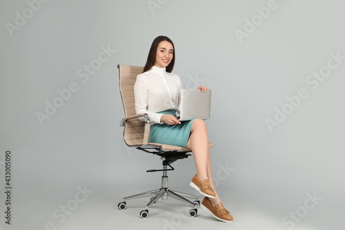 Young businesswoman with laptop sitting in comfortable office chair on grey background