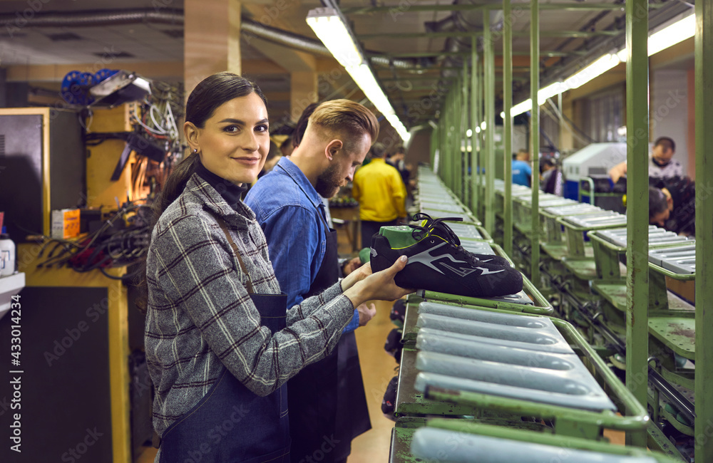 Shoe production manufactory concept. Male and female shoemaker team working at production line at footwear factory. Focus on young woman worker in uniform smiling looking at camera sorting boots