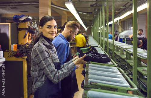 Shoe production manufactory concept. Male and female shoemaker team working at production line at footwear factory. Focus on young woman worker in uniform smiling looking at camera sorting boots © Studio Romantic
