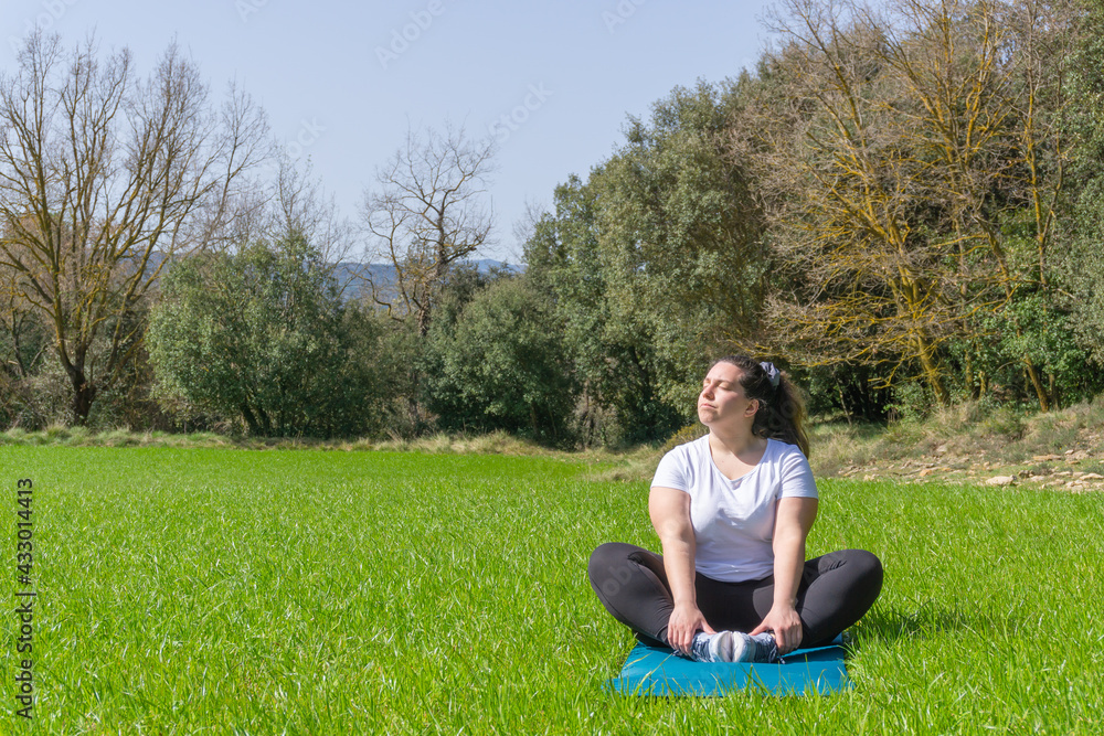 Curvy woman practicing yoga on a mat in the grass looking up at the sun