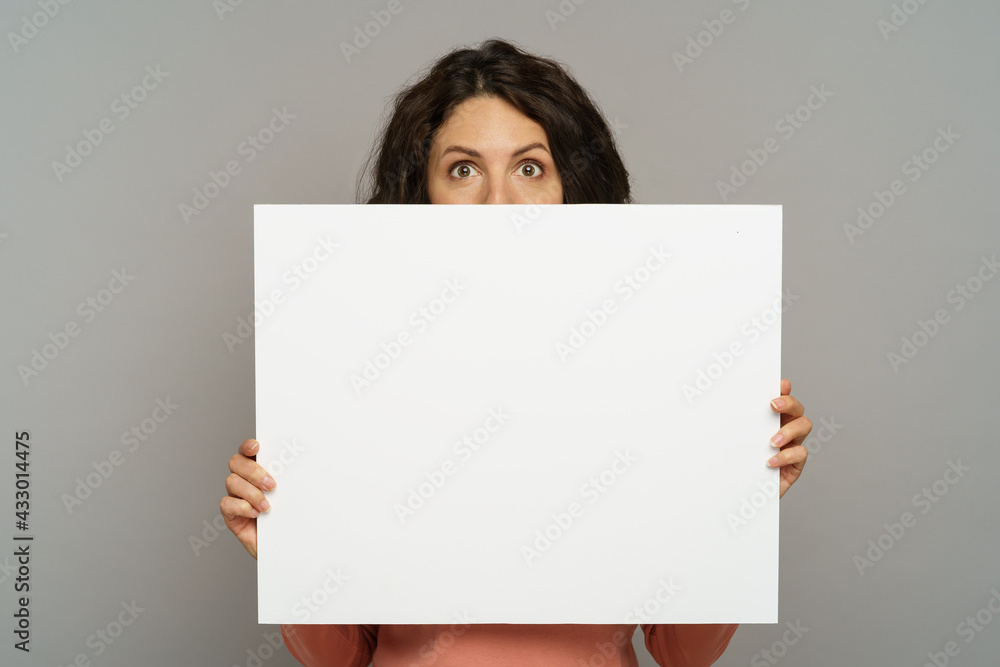 Funny young woman holding empty billboard isolated on grey studio wall background. Joyful brunette millennial girl show white blank poster or banner for business advertisement. Advertising concept Funny young woman holding empty billboard isolated on grey studio wall background. Joyful brunette millennial girl show white blank poster or banner for business advertisement. Advertising concept