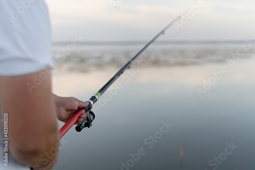 Fishing rod with a float in male hands. The man is a fisherman. River or estuary. Modern fishing tackle. I'm waiting for the coolness. The horizon line is visible in the distance.