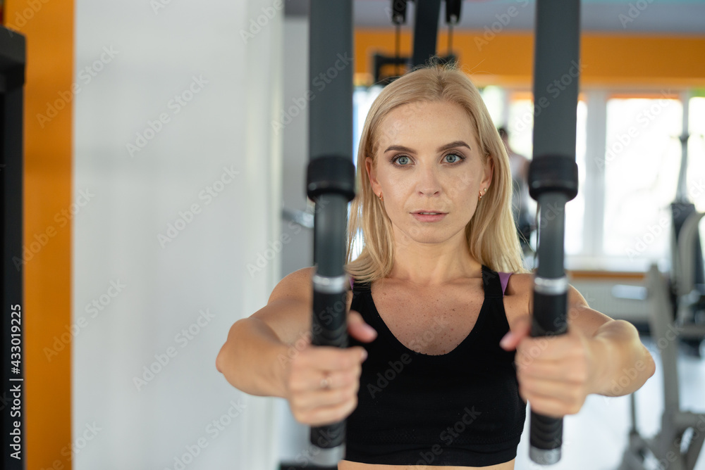 Obraz premium Portrait of the blonde sportswoman and the reduction of hands in the simulator exercise and training of the woman athlete in the gym. Fit muscular girl sitting on a bench pumps up her body.