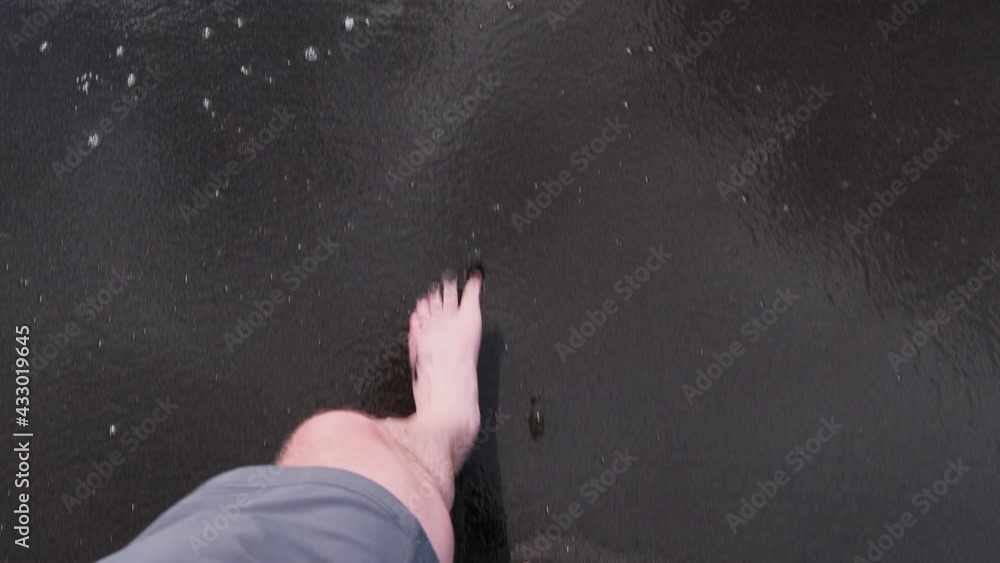 Slow motion top down of barefoot male person walking on black sand beach with splashing water Slow motion top down of barefoot male person walking on black sand beach with splashing water