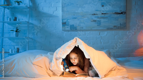 Child under blanker reading book and holding flashlight photo