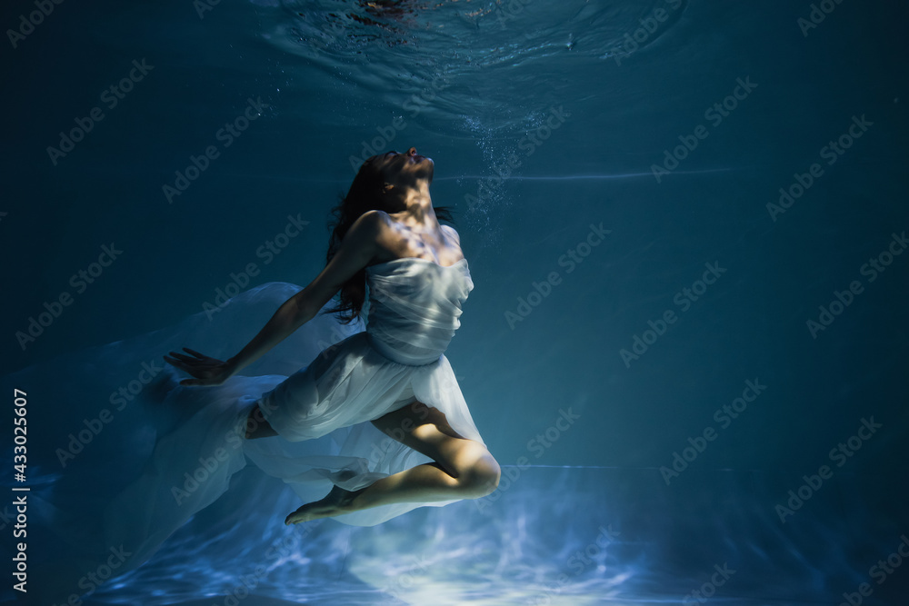 lighting on young graceful woman in white elegant dress swimming in pool with blue water