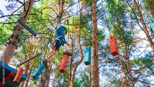 A brave boy moves along a thin rope stretched between trees in a forest rope park. Family outdoor activities with children. Ideas for fun sports activities for children during the summer holidays.