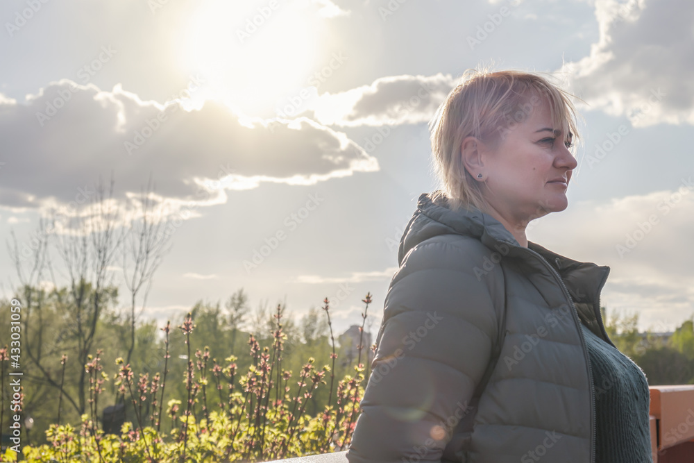 Blonde middle-aged woman in profile stands against the background of the bright setting sun and looks sadly to the side. Concern and heavy thoughts about the future.
