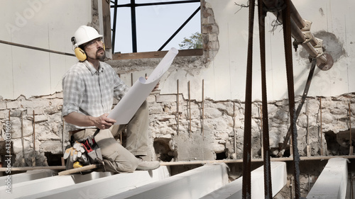 man at work, construction worker wearing helmet and looking at blueprint , check the house project plan, in renovation building site background with the beams at the base of the foundations