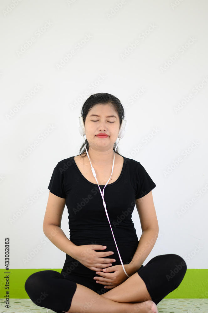 A beautiful pregnant woman wearing headphones with a smile and closing her eyes while sitting on a white background. A beautiful pregnant woman wearing headphones with a smile and closing her eyes while sitting on a white background.