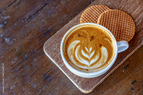 Selective focus a cup of cappuccino, Latte art coffee in white cup on wooden board served with two pieces Dutch waffle (Stroopwafel) Free copy space for your text.