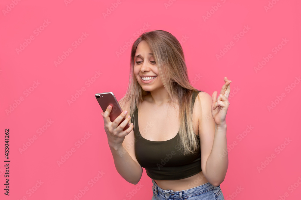 A young pretty caucasian smiling blonde woman crosses her fingers for good luck looking at the phone screen waiting for the results of the lottery or exams isolated on a bright color pink background A young pretty caucasian smiling blonde woman crosses her fingers for good luck looking at the phone screen waiting for the results of the lottery or exams isolated on a bright color pink background