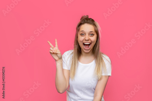 A young pretty caucasian impressed excited smiling cheerful blonde woman in a white t-shirt shows a peace gesture with her hand isolated on a bright color pink background. Girl shows a victory sign