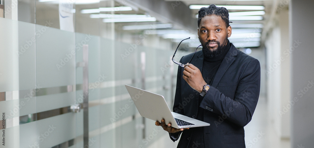 Fototapeta premium Close-up of a dreamy African-American man with glasses, considering new opportunities or an online project strategy, standing with a laptop against the background of a modern office