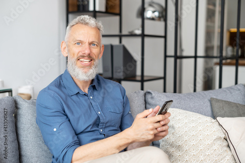 Cheerful and healthy grey-haired middle aged hipster man in casual shirt using smartphone sitting on the sofa at home, mature guy enjoys online chatting, looks at the camera and smiles