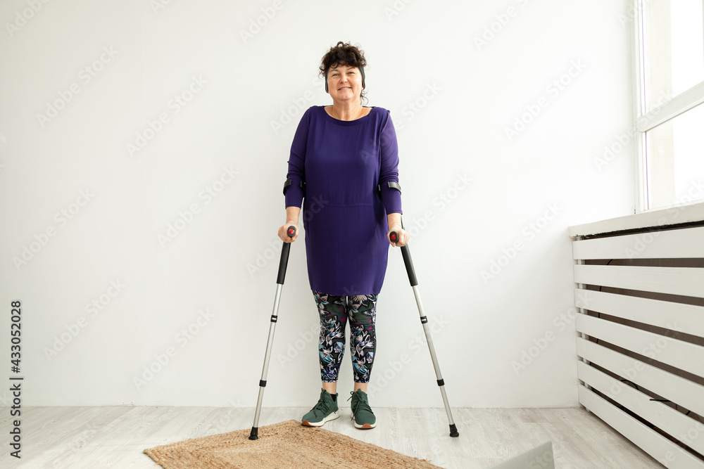 Woman aged stands on white background on crutches in the office of physiotherapist. Trauma recovery and rehabilitation concept. Woman aged stands on white background on crutches in the office of physiotherapist. Trauma recovery and rehabilitation concept.