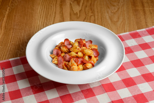 Plate of penne pasta with tomato sauce and meat sausages. Served in a white plate over red plaid table cloth.