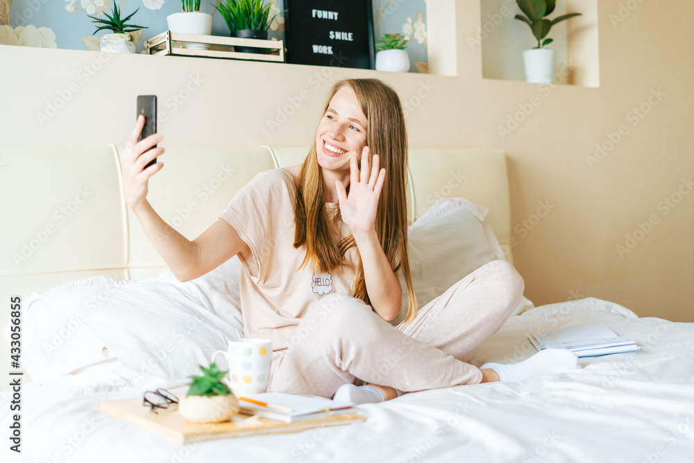 Happy, smiling, relaxed, and cheerful young woman taking a selfie on mobile phone, sitting on bed with coffee. Blonde girl uses cellphone and having video chat. Enjoying free leisure time at home.