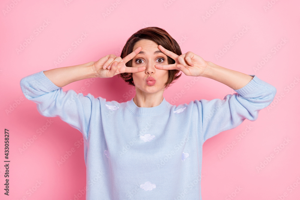 Photo of adorable positive girl kiss lips fingers show v-sign near eyes wear sweater isolated on pink color background Photo of adorable positive girl kiss lips fingers show v-sign near eyes wear sweater isolated on pink color background