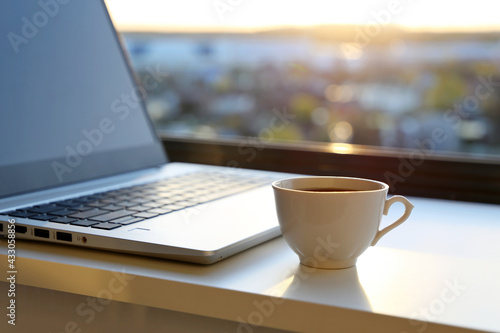 Cozy workplace in home office with hot coffee cup and laptop on table against the window at sunrise. Good morning, concept of remote work and earning online