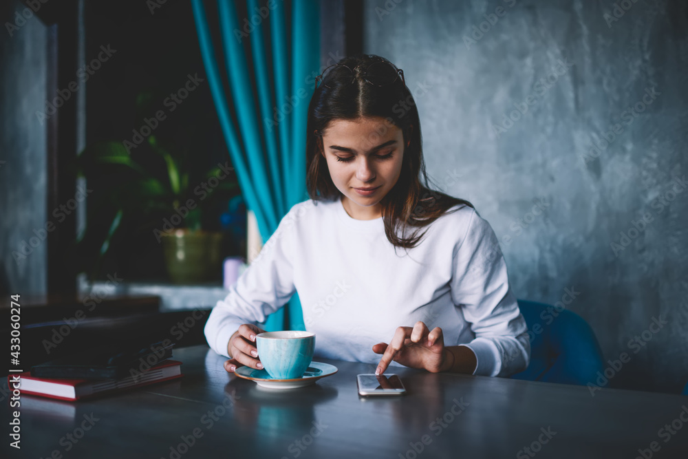 Fototapeta premium Young woman browsing smartphone and having hot beverage in cafeteria