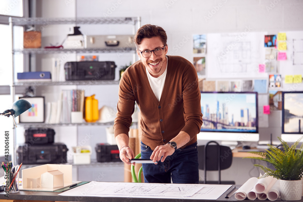 Portrait Of Male Architect At Desk In Office Taking Photo Of Building Plans With Mobile Phone Portrait Of Male Architect At Desk In Office Taking Photo Of Building Plans With Mobile Phone