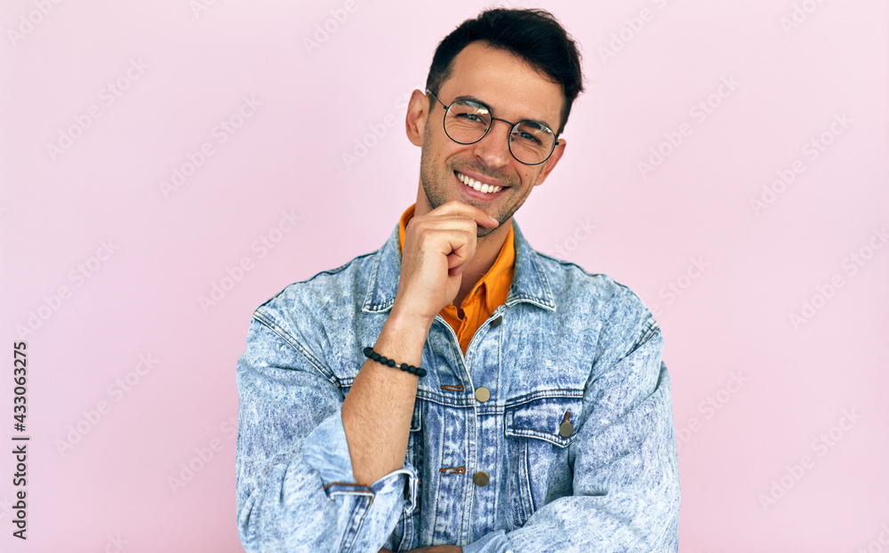Fototapeta premium Positive young man thinking an idea posing over isolated pink background. Male wearing stylish casual clothes, transparent eyeglasses looking to the camera, smiling with a hand on the chin.