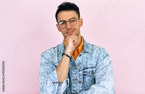 Studio portrait of a thoughtful young man thinking an idea posing over isolated pink background. Male wearing stylish casual clothes, transparent eyeglasses has thoughtful expression.