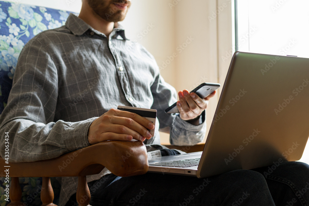 Young man sitting in armchair while shopping online with his credit card, smartphone and laptop from the comfort of his home. Online shopping concept. Young man sitting in armchair while shopping online with his credit card, smartphone and laptop from the comfort of his home. Online shopping concept.