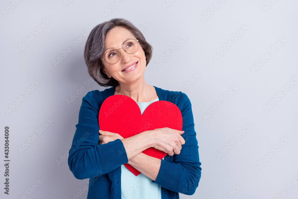 Photo of dreamy charming happy old woman look empty space embrace heart shape isolated on grey color background Photo of dreamy charming happy old woman look empty space embrace heart shape isolated on grey color background