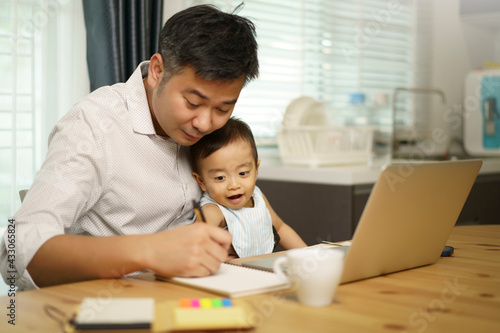 Work from home. Asian young father write notebook and holding his son on knee. looking at notebook. Entrepreneur man working with laptop. A boy sitting with dad at home in kitchen.